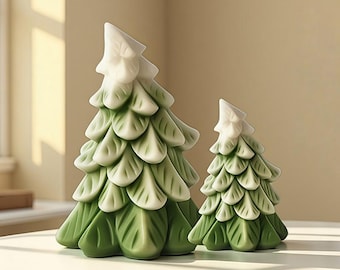 Puede incluir: Dos árboles de Navidad decorativos de cerámica. Los árboles son verdes con puntas blancas y tienen un diseño en capas en forma de hojas. El árbol más grande mide aproximadamente 20 cm de alto, y el más pequeño, unos 13 cm. Están colocados sobre una superficie blanca.