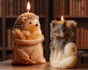 Può includere: Due candele decorative su una superficie di legno. Una candela è a forma di riccio che tiene il grano in un sacco di iuta. L'altra candela è un disegno di volpe grigia e bianca. Entrambe le candele sono accese con una fiamma.