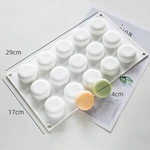 May include: White silicone mold with 18 round cavities, measuring 17 inches by 29 inches. The mold is designed for making small round cakes or other treats. Two small round cakes are shown next to the mold, measuring 4 inches in diameter.