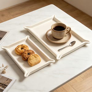Puede incluir: Dos bandejas rectangulares de cerámica color hueso con esquinas elevadas y acampanadas. Una bandeja contiene una taza de cerámica marrón moteada con café negro sobre un plato a juego con una cuchara plateada. La otra contiene tres galletas doradas.