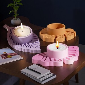 Op de afbeelding: Drie decoratieve kaarshouders in de vorm van bladeren en een bloem. De kaarshouders zijn in tinten lavendel, roze en oranje. Elke houder heeft een brandende kaars. De houders staan op een donkerbruine tafel.