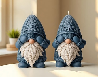 May include: Two blue gnome-shaped candles with white beards and pink noses. The gnomes wear knitted hats with snowflake patterns and matching mittens. The candles are a decorative item.