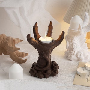 Może przedstawiać: Brązowy, trójpalczasty, szkieletowy świecznik w kształcie dłoni z małą, białą świeczką typu tealight, która się pali w środku. Świecznik stoi na białej powierzchni.