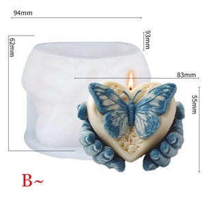 May include: A heart-shaped candle with a blue butterfly design, held by two blue hands. The candle measures 3.3 inches wide and 2.2 inches high. The candle is lit with a small flame. The mold is 3.7 inches wide and 2.4 inches high.