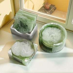 May include: A set of green and white resin coasters and a matching holder. The coasters have a marbled appearance and are square-shaped. A round coaster holder is also included, along with a decorative square coaster.