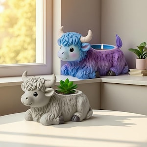 Puede incluir: Dos macetas decorativas con forma de yak. Una es gris y contiene una pequeña suculenta. La otra es azul y morada, con una vela dentro. Ambas tienen cuernos y están sentadas. Las macetas están sobre una superficie blanca.