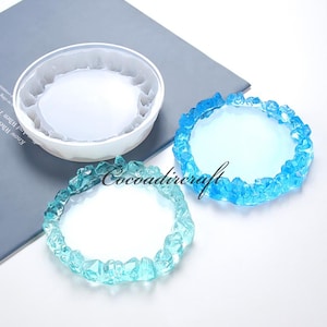 Może przedstawiać: Dwa okrągłe silikonowe formy do rękodzieła z żywicy. Formy są zaprojektowane do tworzenia podkładek pod kubki z krawędzią przypominającą kryształ. Jedna forma jest biała, a druga wypełniona niebieską żywicą.