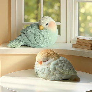 Könnte beinhalten: Zwei Keramikvogel-Figuren, eine auf einer Fensterbank und die andere auf einem Tisch. Die Vögel haben sanfte Pastellfarben, mit Federdetails und geschlossenen Augen. Der Hintergrund zeigt ein Fenster und Bücher. Die Figuren sind dekorative Artikel.