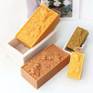 May include: Three rectangular beeswax candles with a bee and flower design. The candles are in different sizes and are displayed on a white surface.