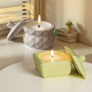 May include: Two lit candles in geometric containers. One is grey with a faceted design, the other is a light green square. Both have lids. A small potted plant and books are in the background.