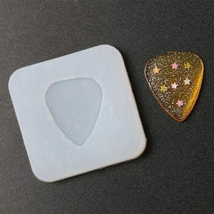 Puede incluir: Un molde de silicona blanco para hacer púas de guitarra. El molde tiene la forma de una púa de guitarra y tiene una superficie lisa. Una púa de guitarra transparente con purpurina y estrellas se muestra junto al molde.