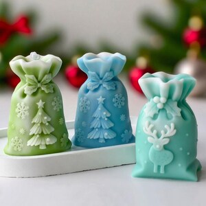 May include: Three festive Christmas-themed bags in green, blue, and teal. Each bag has embossed holiday designs, including snowflakes, Christmas trees, and a reindeer. The bags are displayed on a white tray.