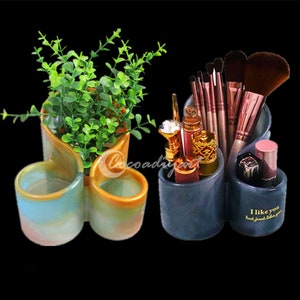 May include: Two ceramic organizers, one in blue and gray with the text "I like you but just like you" and the other in yellow and blue with a plant inside. Both organizers have multiple compartments for holding makeup brushes, lipsticks, and other items.