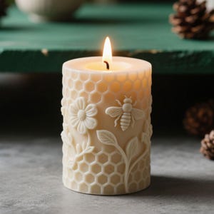 May include: A lit, ivory-colored pillar candle with a honeycomb pattern and embossed floral and bee designs. The candle's surface features a flower, a bee, and leaves. The candle is cylindrical and has a warm, inviting glow.