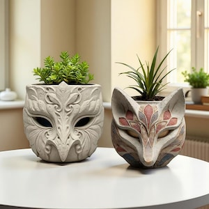 May include: Two decorative planters on a white table. One is a light gray owl mask design, the other a fox face with red, pink, and blue accents. Both planters hold green plants, adding a touch of nature to the decor.