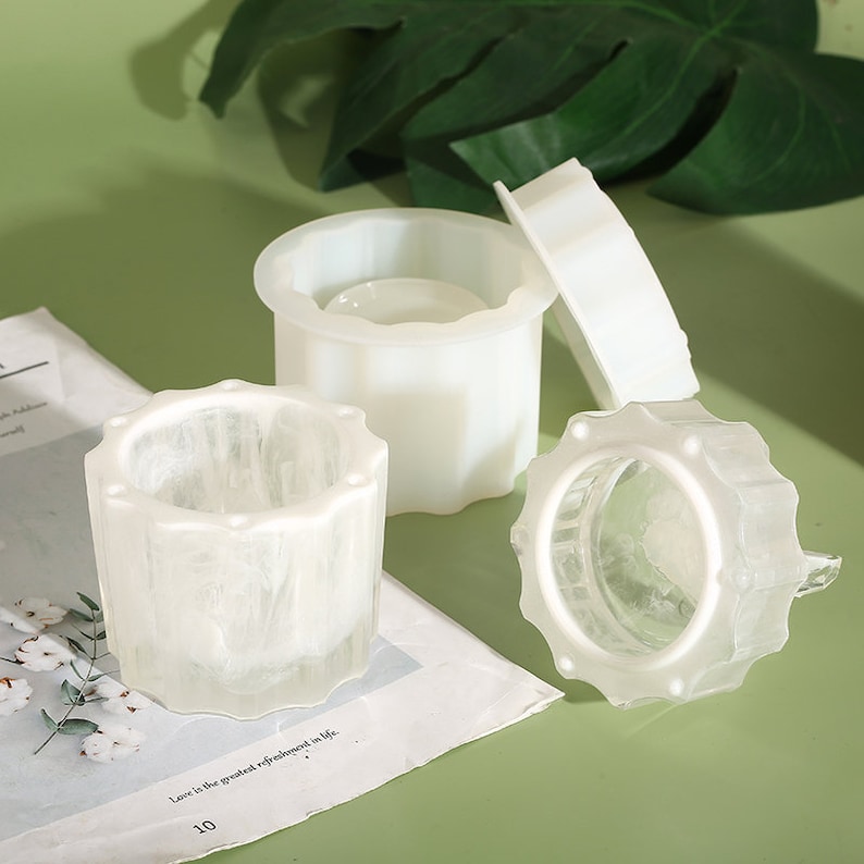 May include: Three white silicone molds for making candles. The molds are in different shapes, including a round mold with a raised rim, a cylindrical mold with a textured surface, and a cylindrical mold with a smooth surface.