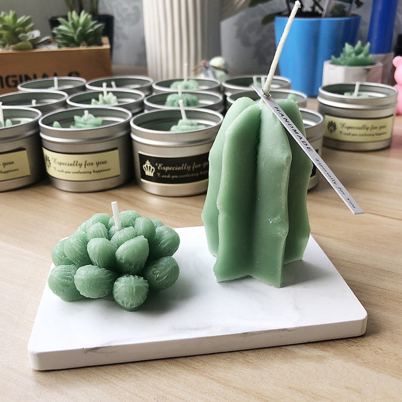 Pode incluir: Duas velas verdes em forma de suculentas em uma bandeja de m&aacute;rmore branco. A vela maior tem a forma de um cacto e tem uma etiqueta que diz "Handmade". A vela menor tem a forma de uma suculenta.