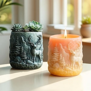 Puede incluir: Dos elementos decorativos: una maceta de color verde azulado con suculentas y una vela. La maceta presenta un diseño de montaña y ciervo. La vela tiene un diseño similar, con una escena de ciervo y montaña, y está encendida.
