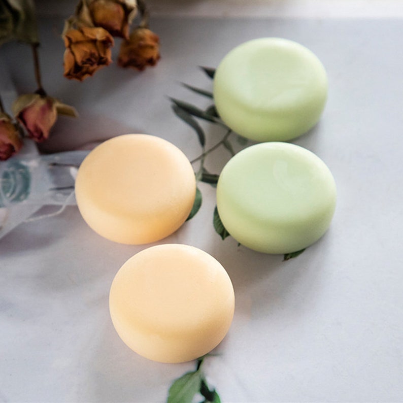 May include: Four round solid shampoo bars, two are light green and two are light orange. The bars are arranged on a white surface.