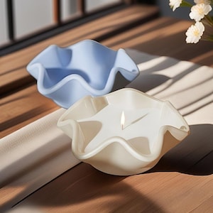 Puede incluir: Dos cuencos decorativos con bordes ondulados, uno azul claro y otro blanquecino, sobre una superficie de madera. El cuenco blanquecino contiene una vela encendida. Los cuencos tienen una forma orgánica única.