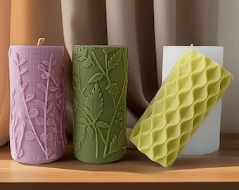 May include: Three decorative pillar candles with different embossed designs. The candle on the left is purple with a floral pattern, the middle candle is green with a leaf pattern, and the candle on the right is yellow with a wavy pattern.