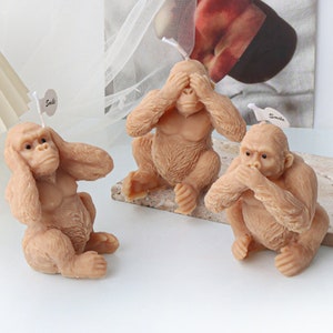 Könnte beinhalten: Drei braune Affen-Kerzenfiguren, jede mit einer anderen Geste, die das Sprichwort "Sehe kein Übel, höre kein Übel, sprich kein Übel" darstellt. Der Affe links hat seine Hände über seinen Ohren, der mittlere Affe hat seine Hände über seinen Augen und der Affe rechts hat seine Hände über seinem Mund.