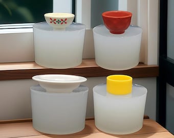 Könnte beinhalten: Vier kleine Schalen in verschiedenen Farben und Designs werden auf transparenten, zylindrischen Sockeln präsentiert. Die Schalen umfassen eine gelbe Schale mit rotem Blumenmuster, eine rote Schale, eine weiße Schale und einen gelben Zylinder.