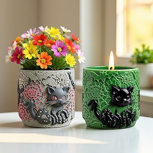 Può includere: Due vasi in ceramica a tema Halloween. Un vaso grigio con un design a gatto e ragnatela contiene fiori colorati. L'altro è un portacandele verde con un design a gatto nero e ragnatela, con una candela accesa.