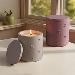 Puede incluir: Dos velas decorativas, una encendida, en un recipiente gris con tapa. La otra es de color malva. Ambas tienen un patrón de cerezas en relieve en los lados. Las velas están sobre una mesa cerca de una ventana.
