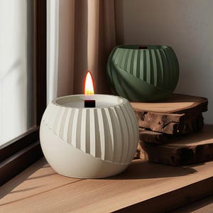 May include: A lit, off-white, spherical candle with vertical grooves and a diagonal accent. A second, unlit, olive-green candle sits behind it. Both candles are on wooden surfaces, near a window. The flame is orange and yellow.