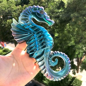 Op de afbeelding: Een iriserend blauw en groen glazen zeepaardje figuur. Het zeepaardje heeft een gedetailleerd ontwerp met schubben en vinnen.