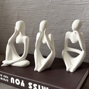Pode incluir: Três esculturas de cerâmica branca de figuras abstratas em posição sentada. As figuras são estilizadas e têm um design minimalista.