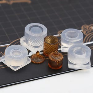 May include: An assortment of clear silicone molds and amber-colored glass containers. The molds are various shapes, including cylindrical and square, with textured and smooth surfaces. The glass pieces include a small jar with a lid and a decorative cup.