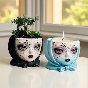 Puede incluir: Dos macetas decorativas con cara de muñeca. Una maceta negra contiene una pequeña planta verde. La otra es un candelabro azul claro con una mecha encendida. Ambas tienen un efecto de porcelana agrietada y están atadas con un lazo.