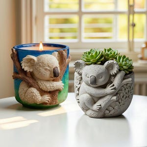 Può includere: Un portacandele blu e verde con un design a koala e una fioriera grigia a forma di koala con piante grasse verdi. Il portacandele ha una candela accesa all'interno. Entrambi gli articoli presentano un koala che abbraccia un ramo d'albero.