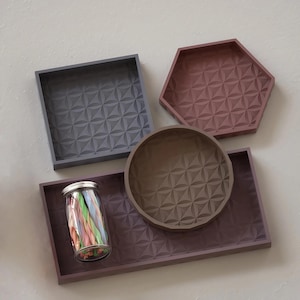 May include: A collection of four trays in square, hexagonal, circular, and rectangular shapes, each featuring a geometric embossed pattern. The trays are coloured in shades of grey, mauve, and brown. A clear glass jar filled with colourful sticks sits inside.