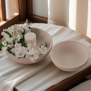 Könnte beinhalten: Zwei hellrosa dekorative Schalen, eine gefüllt mit weißen Blumen und einer Kerze. Die Schalen haben ein geripptes Design an den Rändern. Die Schalen stehen auf einer weißen Tischdecke.