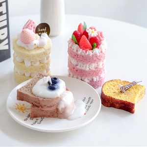 May include: Three decorative candles shaped like cakes. The first cake is a layered vanilla cake with pink frosting and strawberries. The second cake is a layered pink cake with white frosting and strawberries. The third cake is a slice of chocolate cake with white frosting and blueberries. The candles are on a white surface with a white plate.