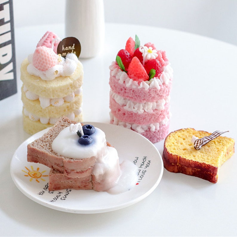 May include: Three decorative candles shaped like cakes. The first cake is a layered vanilla cake with pink frosting and strawberries. The second cake is a layered pink cake with white frosting and strawberries. The third cake is a slice of chocolate cake with white frosting and blueberries. The candles are on a white surface with a white plate.