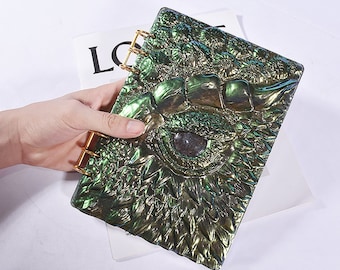 Può includere: Un quaderno verde e iridescente con un designo di occhio di drago sulla copertina. Il quaderno ha anelli dorati ed è chiuso.
