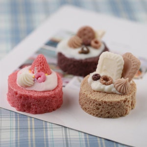 May include: Three miniature cakes decorated with various toppings, including a strawberry, sprinkles, a donut, a cookie, and a chocolate bar. The cakes are on a white surface with a blue and white checkered pattern.