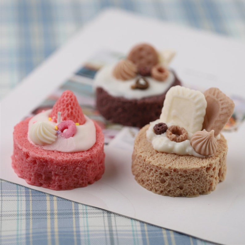 May include: Three miniature cakes decorated with various toppings, including a strawberry, sprinkles, a donut, a cookie, and a chocolate bar. The cakes are on a white surface with a blue and white checkered pattern.