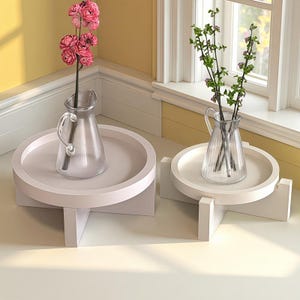 May include: Two white, round, pedestal tables with a light pink hue. Each table holds a clear glass pitcher vase. One vase contains pink flowers, the other has green stems with small purple flowers. The tables are near a window.