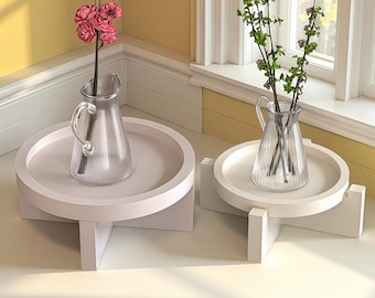 May include: Two white, round, pedestal tables with a light pink hue. Each table holds a clear glass pitcher vase. One vase contains pink flowers, the other has green stems with small purple flowers. The tables are near a window.