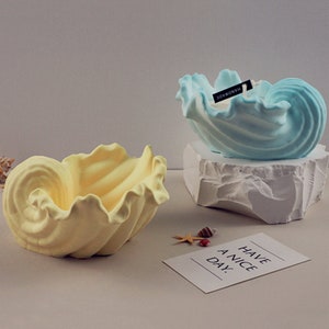 May include: Two decorative seashell-shaped candle holders. One is light blue and the other is yellow. Both are made of ceramic and have a textured surface. The blue candle holder has a small black tag that says "HANDMADE".