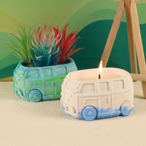 May include: Two white ceramic candles shaped like vintage Volkswagen vans. One van is painted blue and green and has a small potted plant inside. The other van has a lit candle inside.