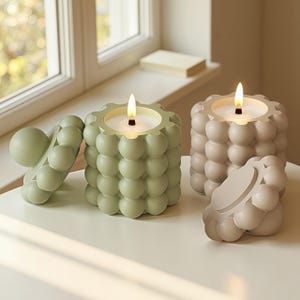 Peut inclure: Deux bougies allumées, à texture de bulles, vert sauge et beige, avec couvercles assortis. Les bougies sont posées sur une surface blanche près d'une fenêtre. Les flammes des bougies sont vives.