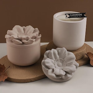 Op de afbeelding: Een verzameling decoratieve kaarsen en een container met deksel. De kaarsen zitten in witte keramische containers, een cilindrische en een ronde met een bloemvormig deksel. De cilindrische kaars heeft een label met de tekst "HANDMADE Especially for You."