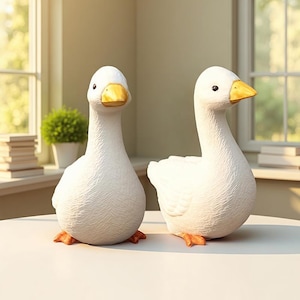 Puede incluir: Dos figuras de gansos blancos con picos amarillos y patas naranjas se exhiben sobre una superficie blanca. Los gansos tienen una superficie texturizada y están colocados frente a una ventana. Una pequeña planta verde y libros son visibles en el fondo.
