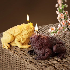 May include: Two frog-shaped candles, one brown and one yellow, with wicks lit. The candles are sitting on a woven wicker surface.